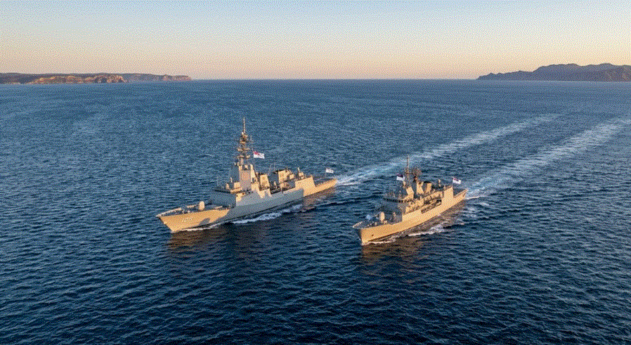قطة جوية لسفينتين حربيتين حديثتين تبحران جنبًا إلى جنب في مياه زرقاء عميقة تحت سماء صافية عند غروب الشمس، مع وجود أرض في الخلفية."