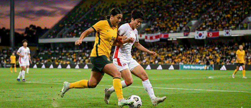 "لقطة حركة قريبة من مباراة كرة قدم نسائية تجمع بين لاعبة أسترالية ترتدي طقماً أصفر ولاعبة كورية شمالية ترتدي طقماً أبيض، وهما تتنافسان على الكرة في ملعب 'بيرث المستطيل' المزدحم خلال وقت الغروب."