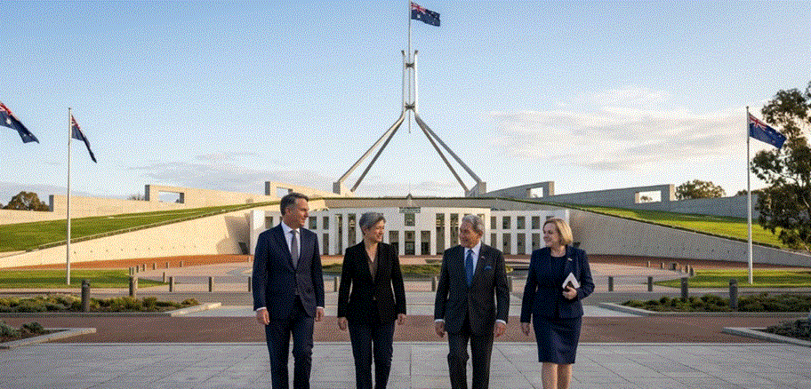 "أربعة مسؤولين حكوميين من أستراليا ونيوزيلندا يسيرون بملابس رسمية أمام مبنى البرلمان الأسترالي في كانبرا، مع وجود أعلام وطنية في الخلفية تحت سماء صافية."