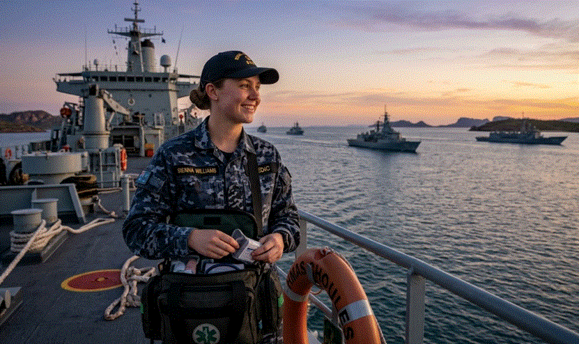 لقطة مقربة منخفضة الزاوية لمسعف بحري أسترالي يرتدي زي التمويه البحري الرسمي (DPCU) أثناء التمرين، ويحمل حقيبة طبية خضراء ويقف أمام خلفية من السفن الحربية والمياه الزرقاء المفتوحة.