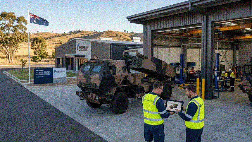 "صورة خارجية أفقية لمنشأة عسكرية أسترالية، تظهر شاحنة راجمة صواريخ هيمارس (HIMARS) متوقفة خارج حظيرة صيانة مفتوحة، وبجانبها مهندسان يرتديان سترات سلامة ويقومان بمراجعة جهاز لوحي."