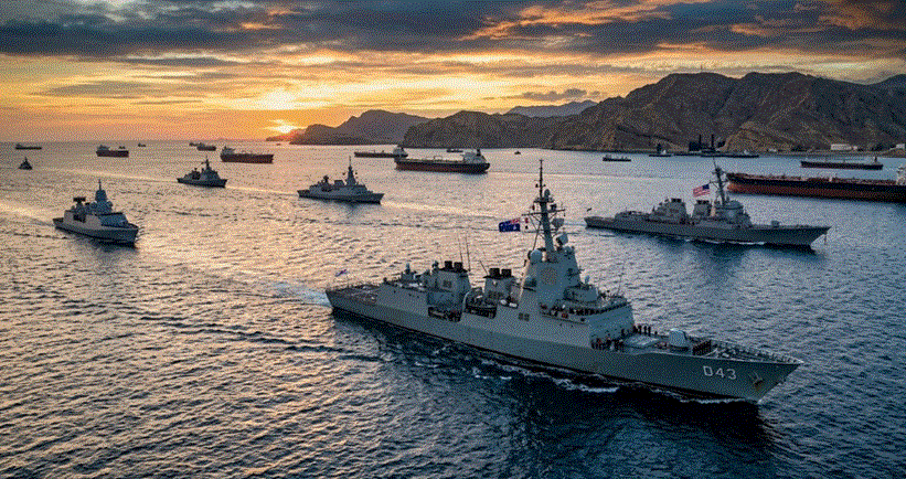 صورة جوية لمضيق هرمز عند الغروب، تظهر مجموعة من السفن الحربية الأسترالية والأمريكية وهي تبحر جنبًا إلى جنب مع ناقلات نفط تجارية، مع وجود ساحل جبلي في الخلفية، مما يبرز الوجود العسكري الدولي لحماية طرق التجارة الحيوية في المنطقة.