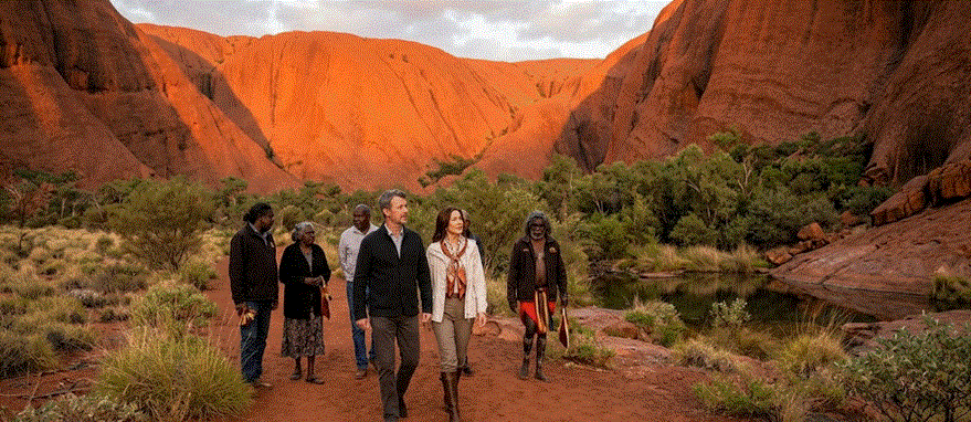 الملك فريدريك والملكة ماري الدنماركيان يسيران في ممر ضيق في صخرة أولورو في أستراليا مع دليل من السكان الأصليين