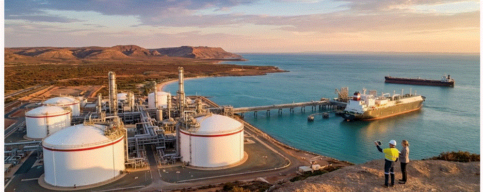 لقطة جوية بانورامية لمصفاة نفط وغاز ساحلية ضخمة في أستراليا أثناء غروب الشمس، تظهر خزانات تخزين بيضاء، وناقلة غاز مسال (LNG) راسية على رصيف بحري، وشخصين يراقبان المشهد من تل مرتفع.