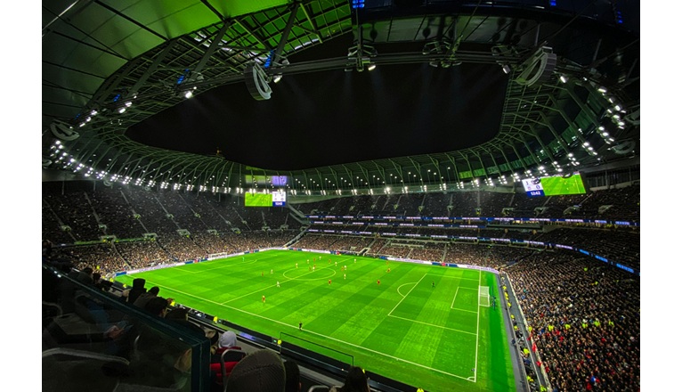 صورة بانورامية التقطت ليلاً لملعب كرة قدم حديث وضخم ممتلئ بالجماهير، تظهر فيه أضواء كاشفة قوية وهيكل سقف مضاء باللون الأخضر، مع شاشتي عرض عملاقتين تظهران تفاصيل المباراة، ومدرجات مزدحمة بالمشجعين يتابعون مباراة جارية على العشب الأخضر.