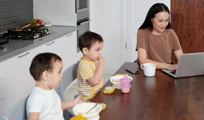 أم تعمل على اللابتوب من المنزل بينما يجلس طفلان صغيران على طاولة الطعام بجانبها