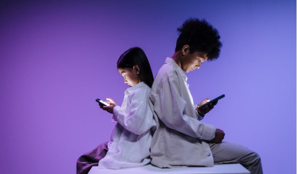 طفلان يجلسان ظهراً لظهر، يستخدمان الهواتف الذكية في إضاءة خافتة، في مشهد يعكس تأثير وسائل التواصل الاجتماعي على الأطفال.