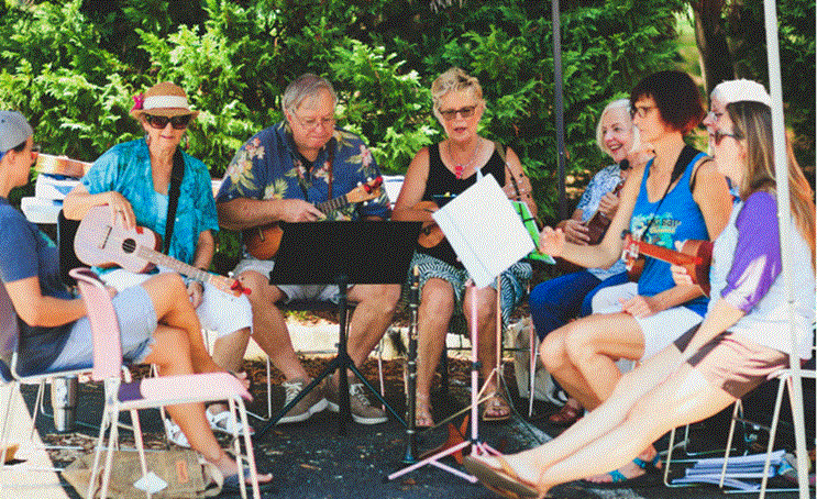 مجموعة من كبار السن يجلسون في الهواء الطلق ويعزفون على آلات اليوكيليلي ويغنون معاً في أجواء اجتماعية مبهجة.