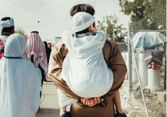 رجل يحمل طفلاً على ظهره وسط مجموعة عائلات في مخيم أو منطقة حدودية، في مشهد يعكس أوضاع اللاجئين والتنقل بين الدول