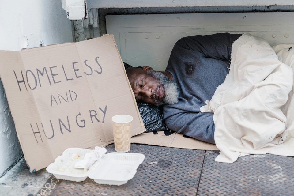 رجل مشرد ينام على الرصيف تحت غطاء أبيض بجوار لافتة كُتب عليها "Homeless and Hungry" في مشهد يعكس أزمة التشرد والفقر في المدن الأسترالية.